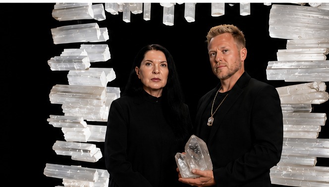 Marina Abramović and Spencer Pratt standing inside a portal of suspended selenite crystals, both dressed in black, with Pratt holding a large clear quartz cluster.